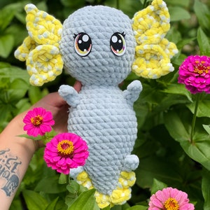 May include: A crocheted gray and yellow axolotl plush toy with large eyes. The toy is sitting in a bed of pink flowers.