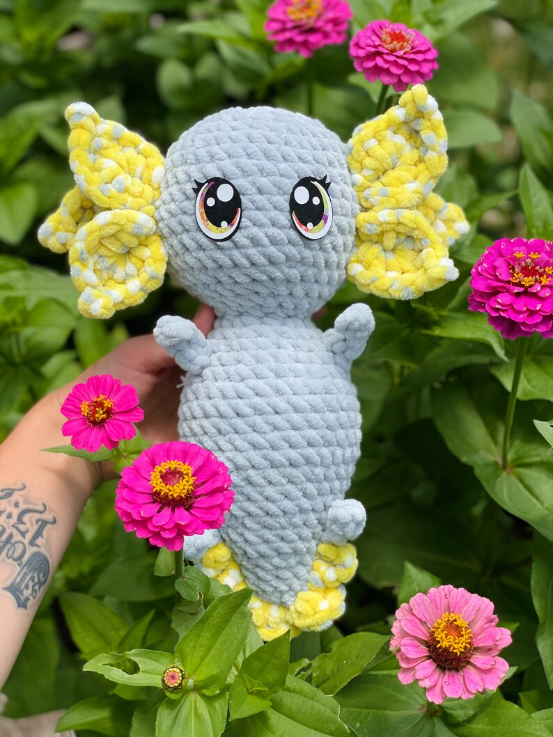 May include: A crocheted gray and yellow axolotl plush toy with large eyes. The toy is sitting in a bed of pink flowers.