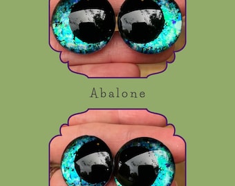 Puede incluir: Dos ojos redondos de vidrio con pupila negra y un patrón de concha de abulón iridiscente. Los ojos son de color azul verdoso con un toque de púrpura.