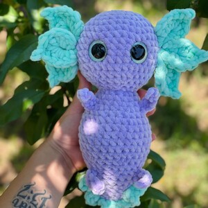 May include: A crocheted axolotl plush toy with purple body and light blue fins. The toy has large black eyes and a detailed crocheted texture.