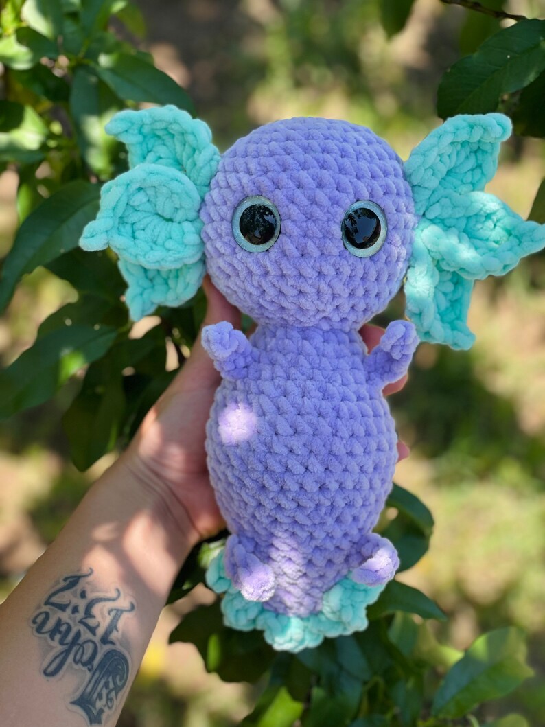 May include: A crocheted axolotl plush toy with purple body and light blue fins. The toy has large black eyes and a detailed crocheted texture.