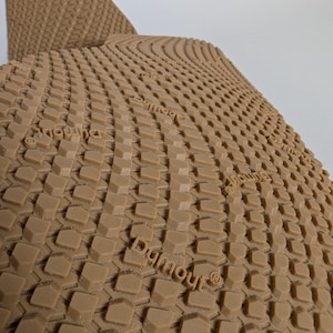 May include: Close-up of a tan shoe sole with a textured pattern of small, raised geometric shapes. The word "Dymour" is imprinted on the sole. The sole is curved, suggesting it is part of a shoe.