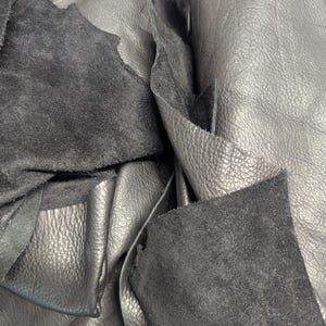 May include: Close-up of black leather scraps with varying textures. Some pieces have a smooth, slightly shiny surface, while others have a rough, suede-like texture. The leather pieces are layered and overlapping, creating a tactile and visually interesting composition.