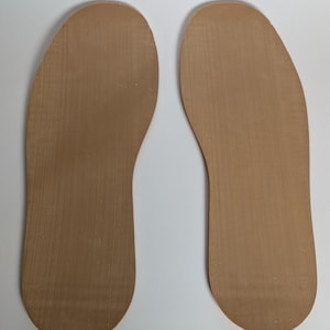 May include: Two tan-colored shoe insoles are displayed against a white background. The insoles are shaped to fit the soles of the feet, with a textured surface. The insoles are made of a flexible material.