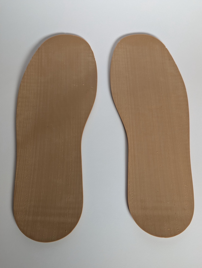 May include: Two tan-colored shoe insoles are displayed against a white background. The insoles are shaped to fit the soles of the feet, with a textured surface. The insoles are made of a flexible material.
