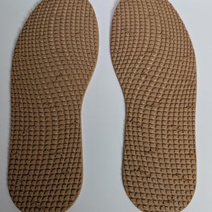 May include: Two brown insoles with a textured, grid-like pattern. The insoles are shaped to fit the foot and have a repeating text pattern. The insoles are designed for comfort and support.