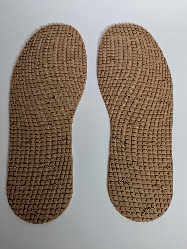 May include: Two brown insoles with a textured, grid-like pattern. The insoles are shaped to fit the foot and have a repeating text pattern. The insoles are designed for comfort and support.