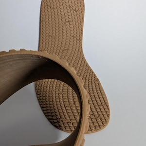 May include: Two brown shoe insoles with a textured pattern are displayed against a white background. One insole is curved, showing the side profile and the textured pattern. The other insole is flat, showing the full shape and pattern.