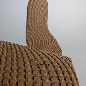 May include: Two brown shoe insoles with textured surfaces. One insole is a full-length shape, the other is a rectangular piece with a repeating geometric pattern. The word "Dymout" is visible on the rectangular piece.