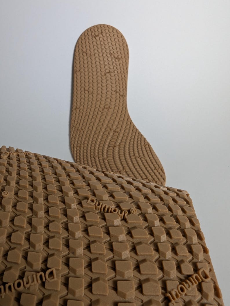 May include: Two brown shoe insoles with textured surfaces. One insole is a full-length shape, the other is a rectangular piece with a repeating geometric pattern. The word "Dymout" is visible on the rectangular piece.