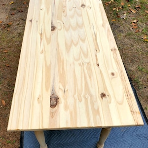Pu&ograve; includere: Un piano da tavolo in legno chiaro con finitura a venatura naturale. Il tavolo ha gambe tornite ed &egrave; appoggiato su un telo blu.