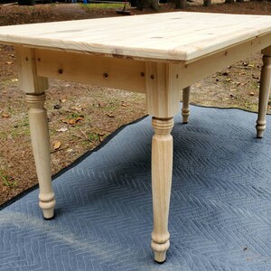 Pu&ograve; includere: Un tavolo in legno chiaro con un piano rettangolare e quattro gambe tornite. Il tavolo &egrave; appoggiato su un tappeto a motivi grigi e neri.