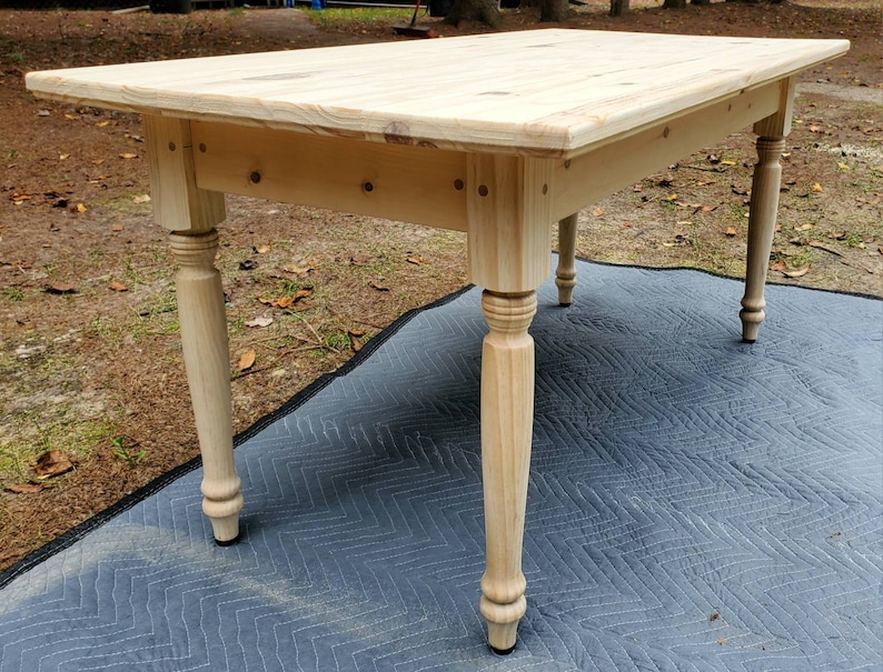 Pu&ograve; includere: Un tavolo in legno chiaro con un piano rettangolare e quattro gambe tornite. Il tavolo &egrave; appoggiato su un tappeto a motivi grigi e neri.