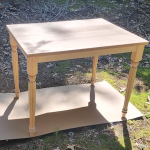 May include: A light-colored wooden table with a rectangular top and four turned legs. The table is approximately 30 inches long, 20 inches wide, and 30 inches high. The table is sitting on a piece of cardboard on the grass.