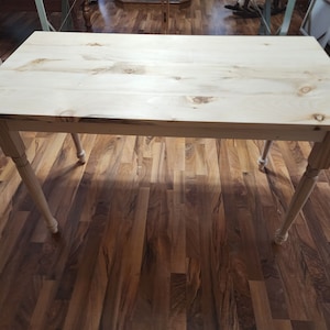 May include: A light wood table with a simple design. The table has a rectangular top and four legs. The legs are turned and have a simple design. The table is made of natural wood and has a light finish.