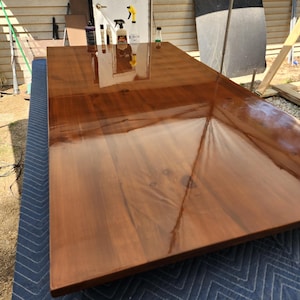 May include: A rectangular wooden tabletop with a smooth, high-gloss finish that reflects the surrounding environment. The wood features a warm, medium-brown tone with visible natural grain patterns and knots throughout the surface.