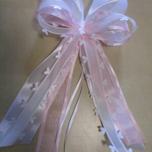 Könnte beinhalten: Eine dekorative Schleife aus weißen und rosa Bändern. Die Schleife hat mehrere Lagen aus Satin- und transparenten Bändern mit kleinen weißen Schmetterlingsakzenten. Geeignet für Geschenkverpackungen oder Eventdekorationen.