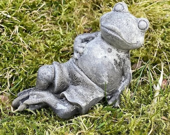 以下が含まれることがあります： リラックスしたポーズで横たわっている、灰色のコンクリート製のカエルの置物。脚は伸ばされ、彫刻されたショートパンツを履いています。置物は緑の苔の上に置かれています。