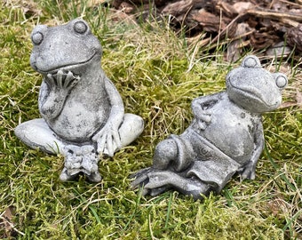 Könnte beinhalten: Zwei graue Beton-Froschfiguren, eine sitzt mit den Händen vor sich gefaltet, die andere liegt auf dem Rücken mit ausgestreckten Beinen.