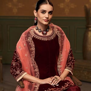Puede incluir: Un salwar kameez de terciopelo granate con bordados dorados y un dupatta de red melocot&oacute;n. El atuendo presenta un escote redondo y mangas tres cuartos. La modelo lleva un collar y pendientes a juego.