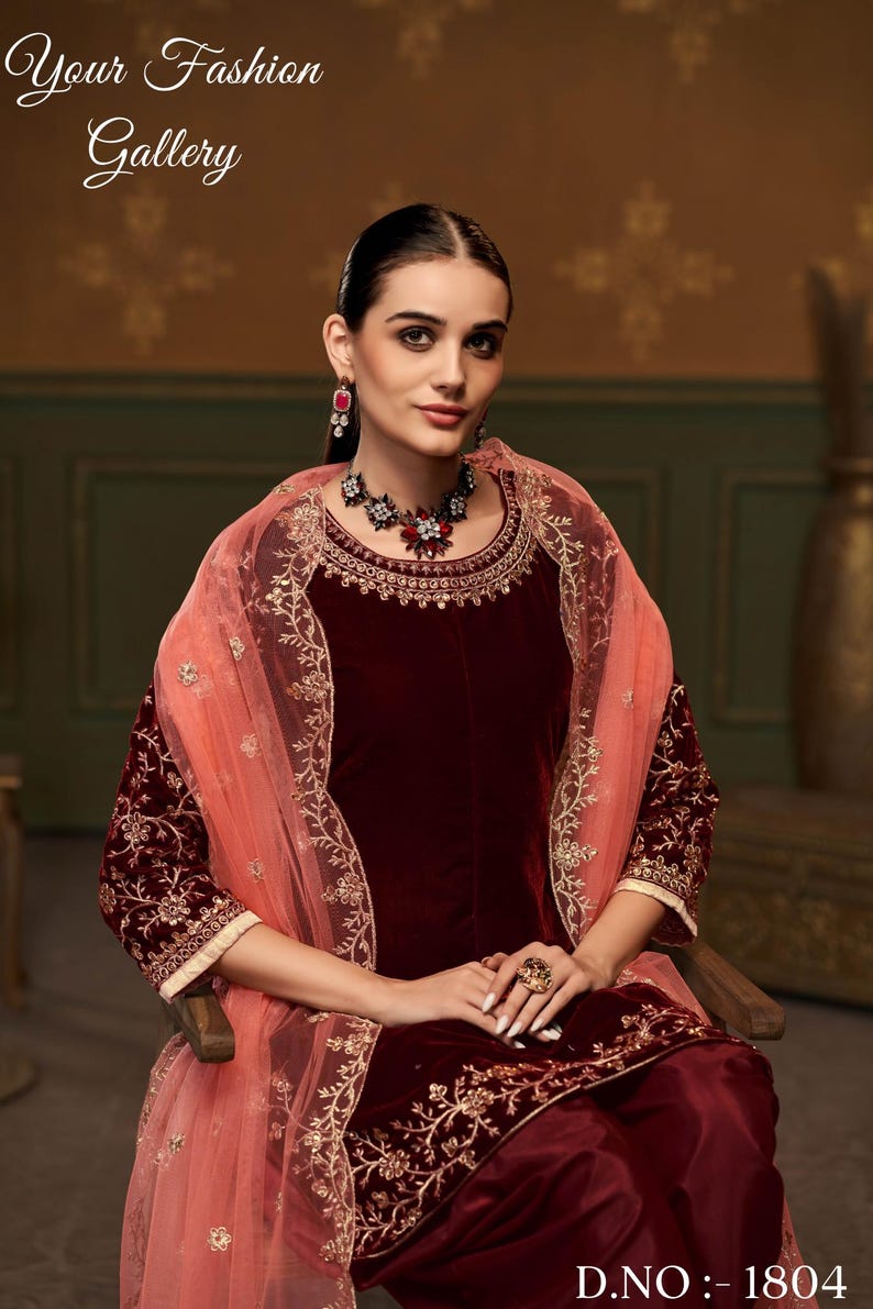 Puede incluir: Un salwar kameez de terciopelo granate con bordados dorados y un dupatta de red melocot&oacute;n. El atuendo presenta un escote redondo y mangas tres cuartos. La modelo lleva un collar y pendientes a juego.