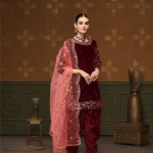 Puede incluir: Un salwar kameez de terciopelo granate con bordados dorados y un dupatta a juego. El dupatta es de color melocot&oacute;n claro y transparente con bordados dorados. El atuendo lo lleva una modelo de pie frente a un fondo marr&oacute;n con estampado. D.NO: 1804