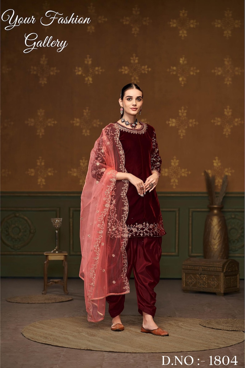 Puede incluir: Un salwar kameez de terciopelo granate con bordados dorados y un dupatta a juego. El dupatta es de color melocot&oacute;n claro y transparente con bordados dorados. El atuendo lo lleva una modelo de pie frente a un fondo marr&oacute;n con estampado. D.NO: 1804