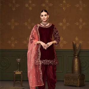 Puede incluir: Un salwar kameez de terciopelo granate con bordados dorados. El kameez tiene un escote redondo y mangas tres cuartos. El salwar es un par de pantalones holgados. El atuendo se combina con un dupatta de red rosa con bordados dorados.