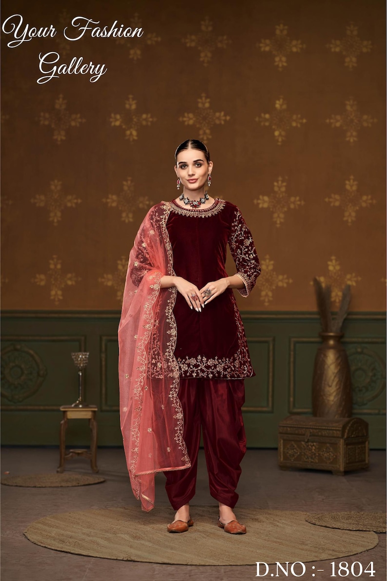 Puede incluir: Un salwar kameez de terciopelo granate con bordados dorados. El kameez tiene un escote redondo y mangas tres cuartos. El salwar es un par de pantalones holgados. El atuendo se combina con un dupatta de red rosa con bordados dorados.