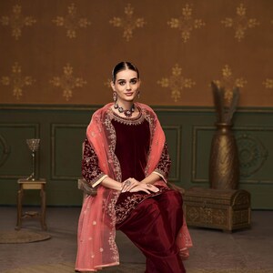 Puede incluir: Una mujer lleva un salwar kameez de terciopelo granate con bordados dorados y un dupatta de red melocot&oacute;n. Est&aacute; sentada en una silla frente a una pared marr&oacute;n con un patr&oacute;n floral. El texto "D.NO: - 1804" es visible en la parte inferior de la imagen.
