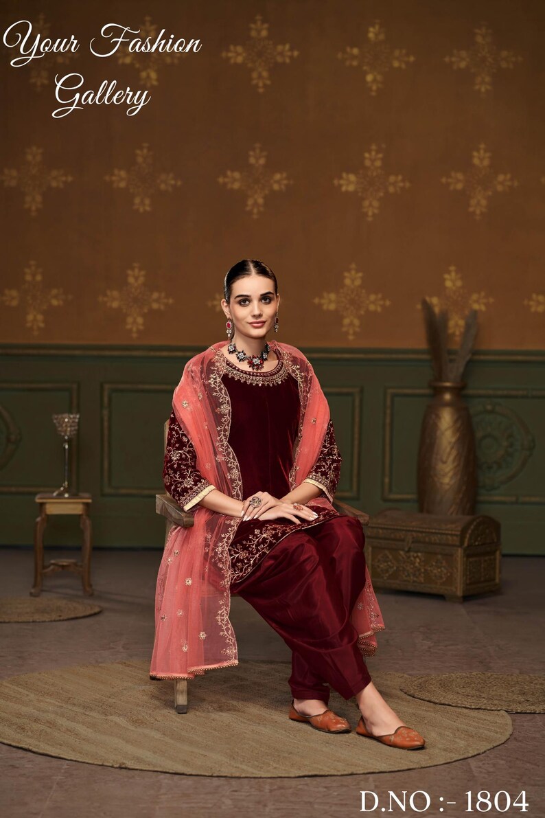 Puede incluir: Una mujer lleva un salwar kameez de terciopelo granate con bordados dorados y un dupatta de red melocot&oacute;n. Est&aacute; sentada en una silla frente a una pared marr&oacute;n con un patr&oacute;n floral. El texto "D.NO: - 1804" es visible en la parte inferior de la imagen.