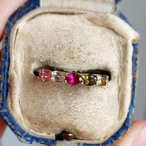 May include: A gold band ring with a row of colorful gemstones. The ring is in a blue velvet ring box.