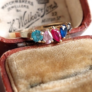 Puede incluir: Un anillo de oro con cinco piedras preciosas de diferentes colores. Las piedras preciosas son azules, rosas, rojas y dos tonos de azul. El anillo está en una pequeña caja de terciopelo rojo.