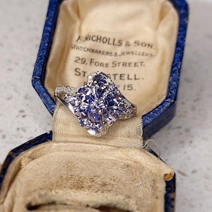 Könnte beinhalten: Ein blauer Saphirring mit einem floralen Design, das in Silber gefasst ist. Der Ring befindet sich in einer blauen Samtringbox mit der Aufschrift "ANICHOLLS & SON. WATCHMAKERS & JEWELLERS 29, FORE STREET. STTELL. 15."