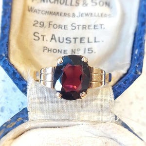 May include: A gold ring with a large, dark red oval gemstone. The ring is in a blue velvet box with a white label that reads "A Nicholls & Son, Watchmakers & Jewellers, 29, Fore Street, St. Austell. Phone No. 15."