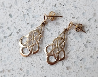 May include: A pair of gold Celtic knot earrings. The earrings are in the shape of a knot with a teardrop shape. The earrings are on a white background.