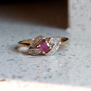 May include: A gold ring with a central oval ruby gemstone. The ring has a leaf-like design on either side of the ruby, with small diamonds. The band is a simple gold colour.