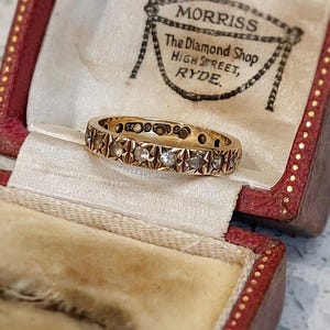 May include: A gold ring set with small diamonds in a star pattern, presented in a red jewellery box. The box features the text "MORRISS The Diamond Shop HIGH STREET, RYDE."