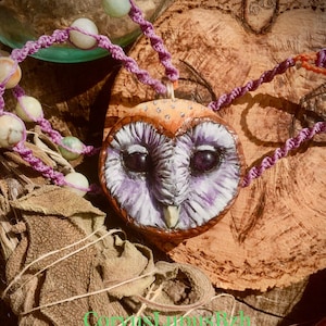 Könnte beinhalten: Eine Halskette mit einem Anhänger in Form einer lila und braunen Eule an einer Perlenkette. Die Eule hat große, dunkle Augen und ein detailliertes Design. Die Halskette liegt auf einem Stück getrocknetem Salbei.