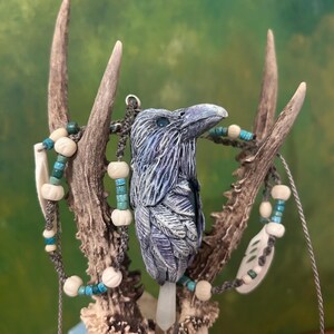 Peut inclure: Un pendentif corbeau composé d'une sculpture d'oiseau détaillée en gris et bleu avec un cristal blanc pointu à la base. Le cordon du collier est orné de petites perles turquoise, de perles sculptées crème et d'éléments blancs en forme d'os.