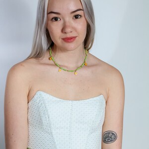 Puede incluir: Una mujer con cabello rubio lleva un top de corsé blanco con ribete verde. También lleva un collar de cuentas verdes y amarillas con pequeños dijes amarillos. Tiene un pequeño tatuaje en el brazo.