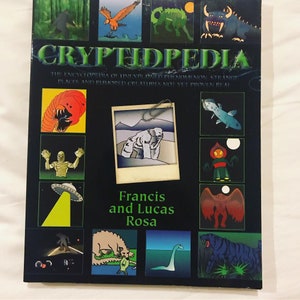 Op de afbeelding: Een boekomslag met een zwarte achtergrond en kleurrijke illustraties van mythische wezens. De titel van het boek is "Cryptopedia: De encyclopedie van onverklaarde fenomenen, vreemde plaatsen en geruchten over wezens die nog niet bewezen zijn". De auteurs zijn Francis en Lucas Rosa.