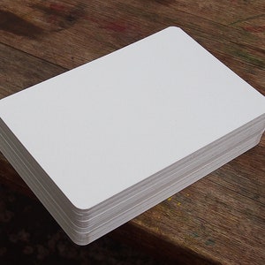 May include: A stack of blank, white rectangular cards with rounded corners. The cards are neatly stacked on a wooden surface, suggesting a stationery or crafting product. The edges of the cards are visible, showing the thickness of the stack.