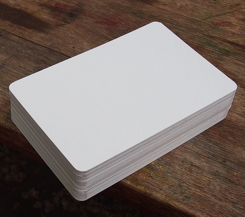 May include: A stack of blank, white rectangular cards with rounded corners. The cards are neatly stacked on a wooden surface, suggesting a stationery or crafting product. The edges of the cards are visible, showing the thickness of the stack.