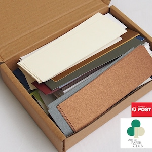 May include: A cardboard box filled with various coloured paper strips. The box is open and the paper strips are visible. The paper strips are in different colours, including brown, green, blue, and red. The box is labelled "Artist Paper Club".