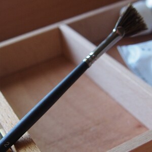 May include: A black handled fan brush with a natural bristle fan. The brush is lying in a wooden box.
