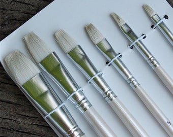 May include: A set of artist paint brushes with natural bristles, metal ferrules, and wooden handles. The brushes are secured with light blue rubber bands and arranged on a white surface. The handles are numbered.