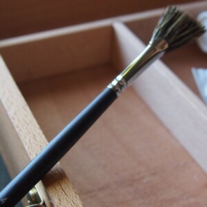 May include: A black handled paint brush with a fan shaped brush head. The brush is in a wooden box.