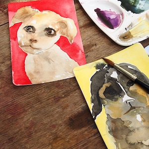 May include: Watercolor paintings of dogs on rectangular cards. One dog is light brown with a red background, the other is black and brown with a yellow background. A paintbrush and a palette with paint are also visible.