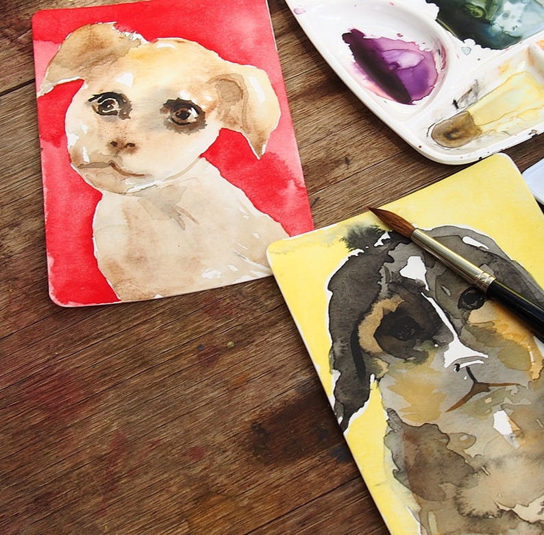 May include: Watercolor paintings of dogs on rectangular cards. One dog is light brown with a red background, the other is black and brown with a yellow background. A paintbrush and a palette with paint are also visible.